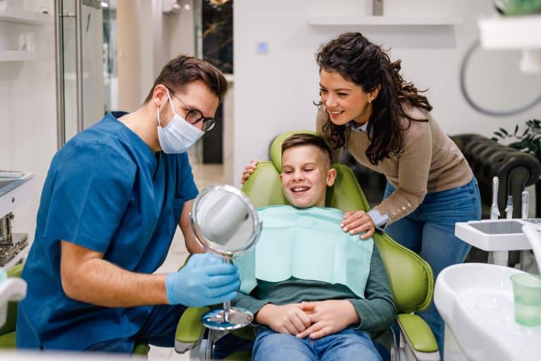 Dentist holds mirror for boy to see his teeth, mother watches contentedly