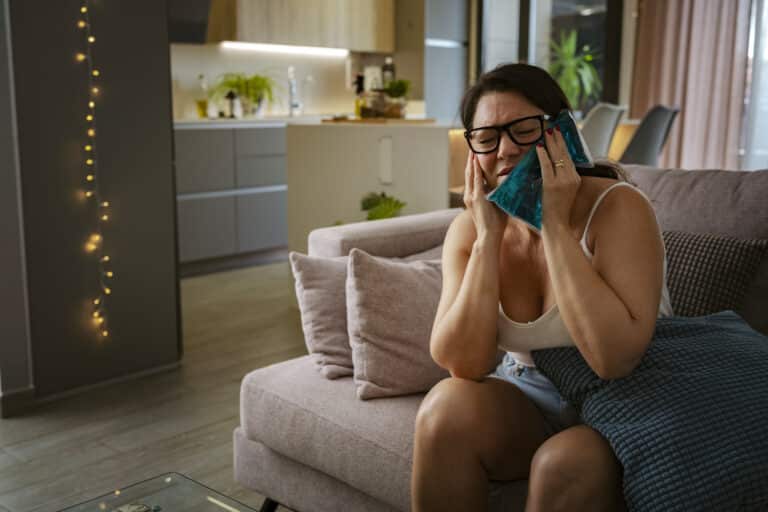 Dental Tooth Pain Woman sitting on the sofa with a severe toothache applying ice gel pack on her jaw. Uncomfortable and displeased, eyes closed person at home